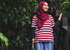 A Muslim woman wearing a hijab stands on wooden stairs in a lush green forest, smiling warmly.