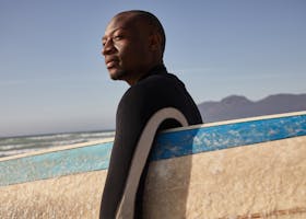 African man holding a surfboard by the sea on a sunny day, ready for surfing.